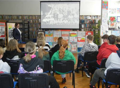 Widok od tyłu na młodzież uczestniczącą w zajęciach w bibliotece, w tle widać prowadzącą dr Barbarę Męczykowską oraz ekran z prezentacją.